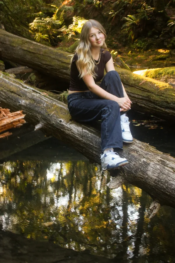 Lynden seated on a fallen log beside the creek in soft forest light
